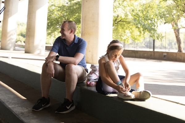 A man and a young girl sit under a shaded area, with the girl tying her shoes after rollerskating..