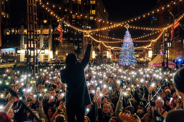 A performer faces a large crowd holding up phone lights during a holiday tree lighting event at The Wharf in Washington, DC.