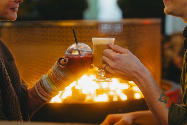 A couple clinks together two festival holiday cocktails in front of a fire.
