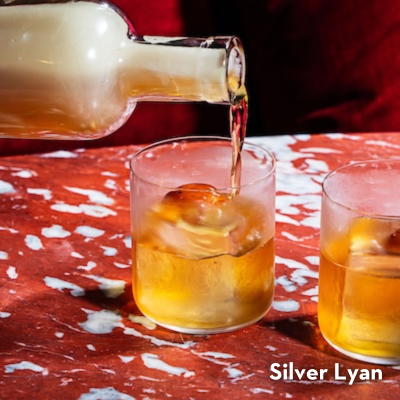 A bottle pours an elegant cocktail into a whiskey glass, against a red marble table. 