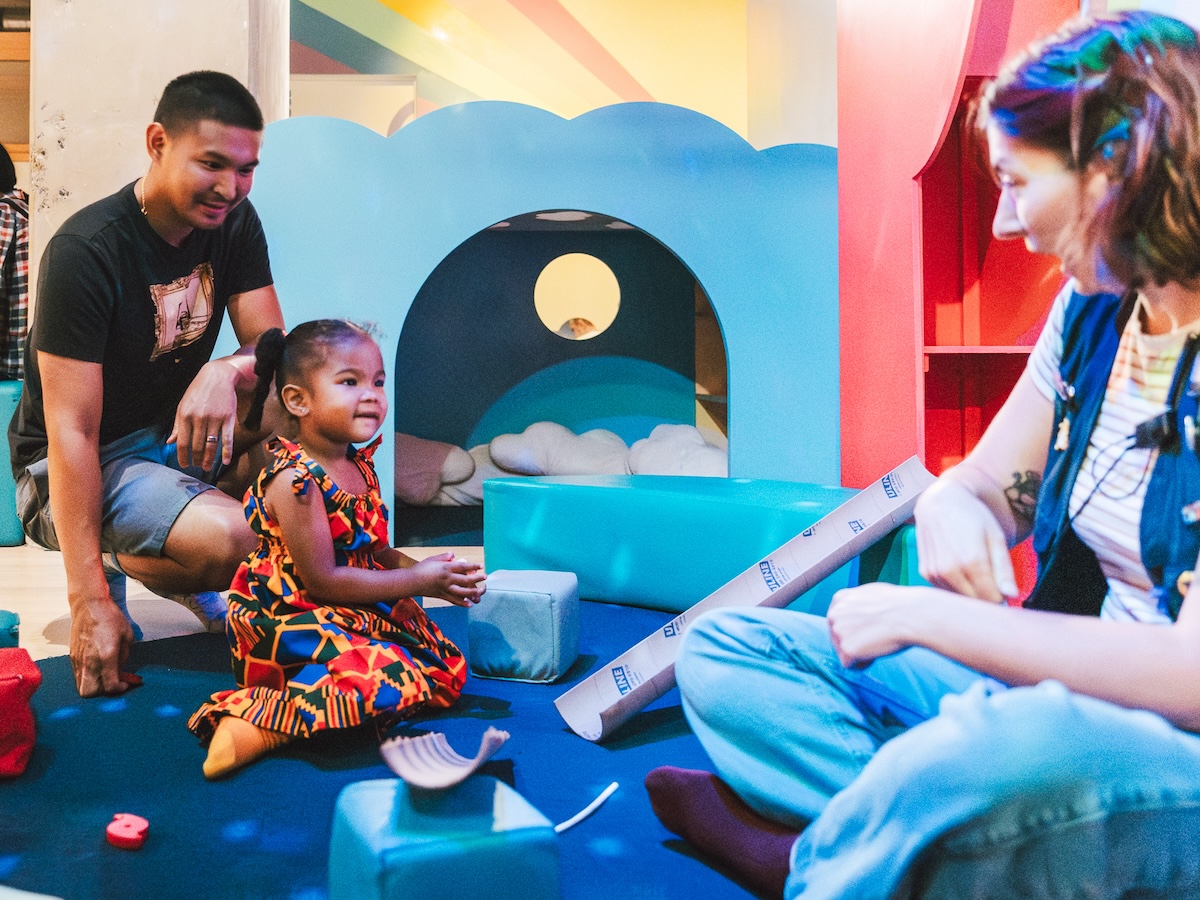 A young child plays with soft blocks in a colorful museum play space while two adults engage with her.