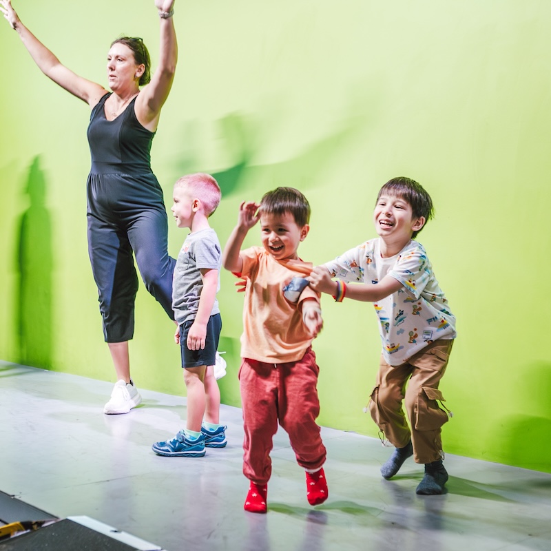 Children laugh and move energetically in a green-screen activity area while an adult leads the group.