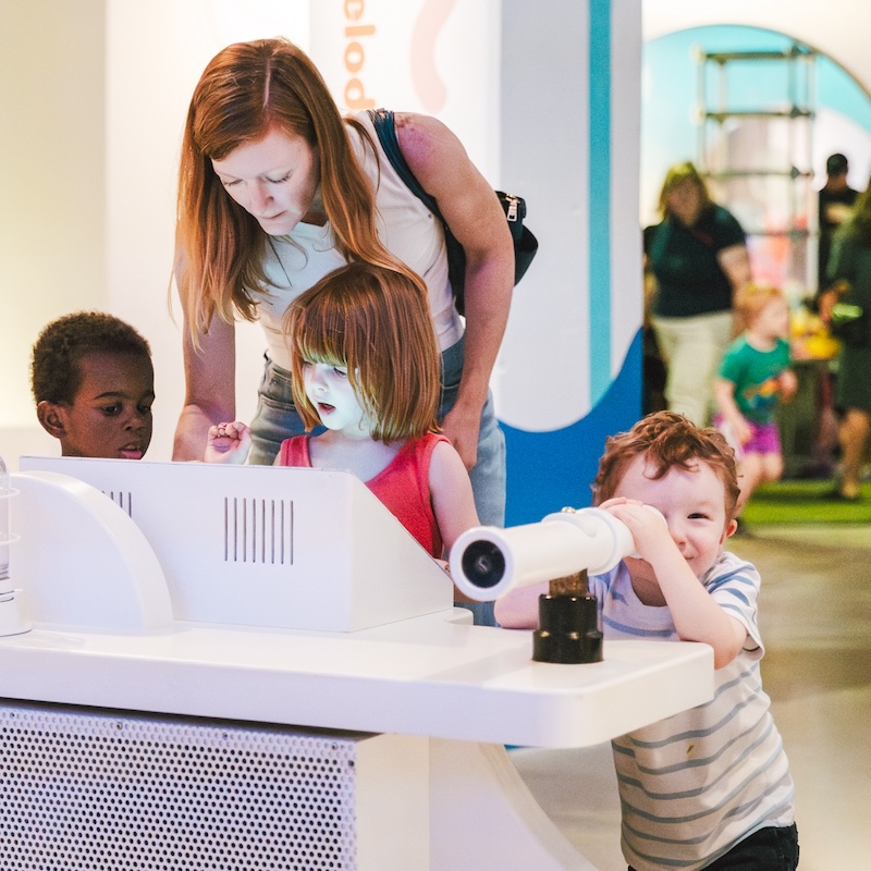 Children explore an interactive station at a museum, with one child peering through a play telescope as an adult helps others at a touchscreen.