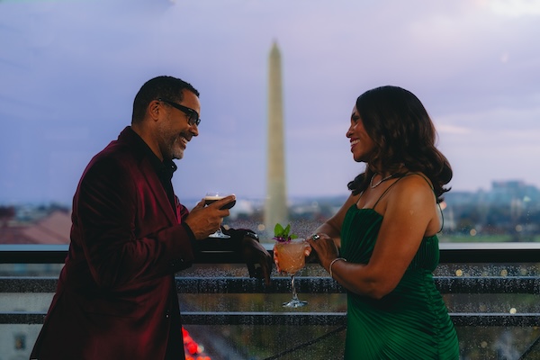 A couple enjoys cocktails on a rooftop terrace with the Washington Monument visible in the background at dusk.
