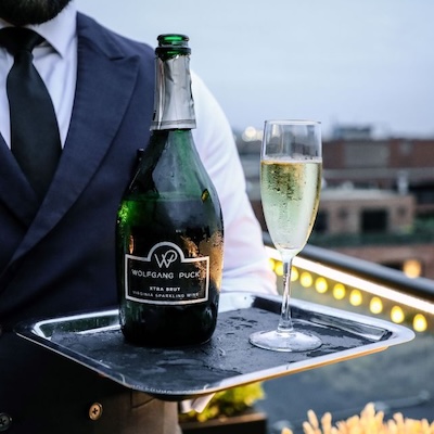 A server presents a tray with a bottle of Wolfgang Puck sparkling wine and a filled champagne glass on a rooftop.