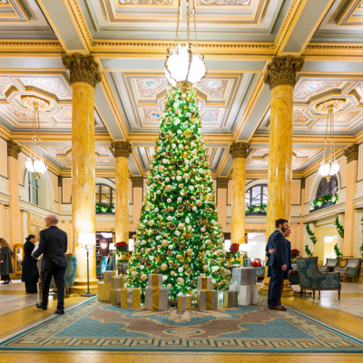 A towering Christmas tree decorated in green, gold, and silver stands in an ornate hotel lobby with guests mingling beneath chandeliers in Washington, DC.