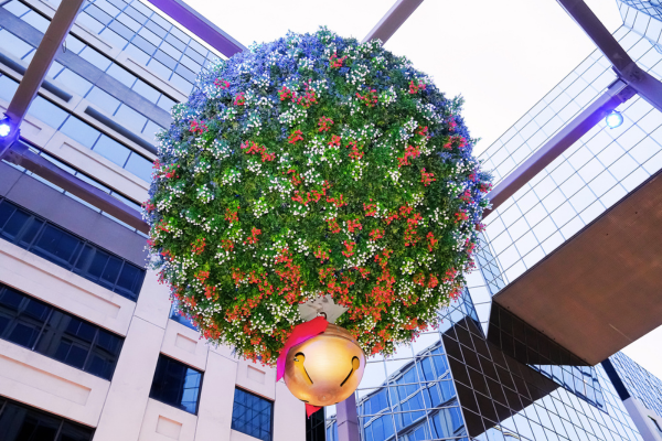 A close-up view of a giant holiday ornament covered in greenery and lights suspended between modern buildings at CityCenterDC in Washington, DC.