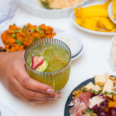 A hand holds a green cocktail garnished with fruit on a table set with small plates and charcuterie in Washington, DC.