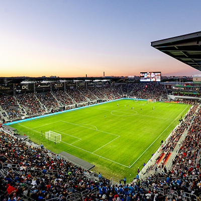 Fans fill Audi Field as a DC United soccer match unfolds under a colorful sunset sky in Washington, DC.
