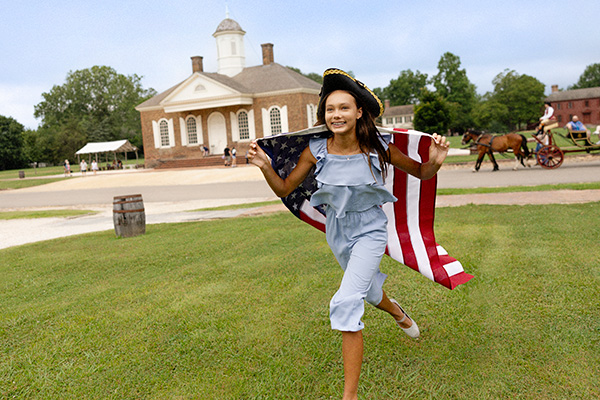 A child in colonial-era attire runs across a historic site lawn holding an American flag in Washington, DC.