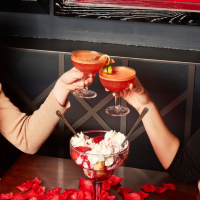 Two friends clink coupe glasses filled with red cocktails over a dessert topped with whipped cream and rose petals at a cozy bar table.