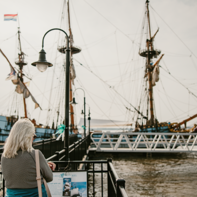 A woman reading an informational plaque in Historic New Castle in New Castle, DE, as the Tall Ship of Delaware, the Kalmar Nyckel, sits ahead of her by a pier.