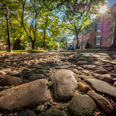 A ground view of a cobblestone street in Historic New Castle in New Castle, DE, with the sun shining through the trees above and historic buildings standing in the distance.