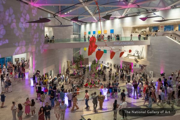 Crowds of visitors gather in the National Gallery of Art’s atrium during NGA Nights, with colorful lighting, suspended art installations and people mingling, dancing and exploring the galleries.
