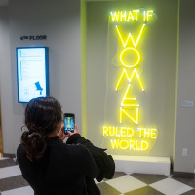 Visitor photographing a bright yellow neon sign reading “What if women ruled the world” on display at the National Museum of Women in the Arts.