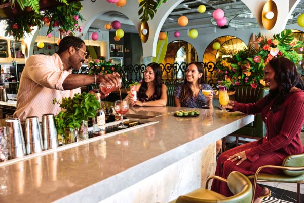Bartender pouring a cocktail behind a marble bar as three women sit on colorful stools, smiling and chatting in a vibrant, plant-filled restaurant with bright hanging decorations overhead.
