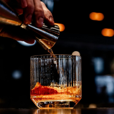 Close-up of a bartender pouring amber-colored liquor from a jigger into a textured rocks glass with a large ice cube on a bar.