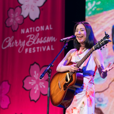 Woman playing an acoustic guitar and singing on stage in front of a pink “National Cherry Blossom Festival” backdrop decorated with cherry blossom graphics.