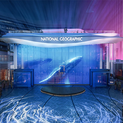 Illuminated entrance to the National Geographic Museum of Exploration at night, with a large glass façade displaying digital ocean imagery and glowing blue light patterns across the plaza.