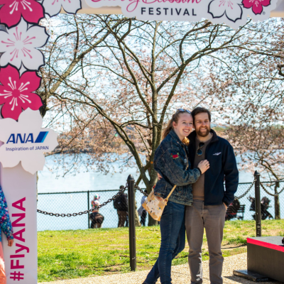Couple posing for a photo at a BloomFest cherry blossom festival frame near the Tidal Basin, with blooming trees and the water in the background.