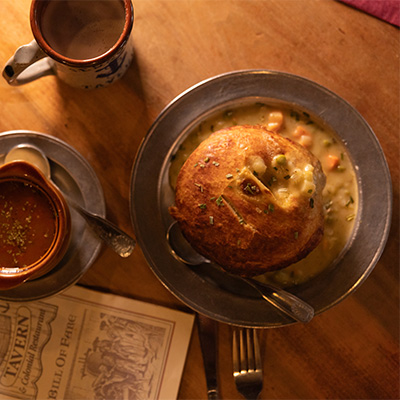 Overhead view of a rustic table setting at Jessop’s Tavern in New Castle, Delaware, featuring a bread bowl filled with creamy soup, a side of broth and a ceramic mug, evoking a warm, colonial-era dining experience.