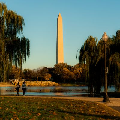 Washington Monument rising above Constitution Gardens at sunset, with willow trees, a pond and people walking along the path in warm golden light.