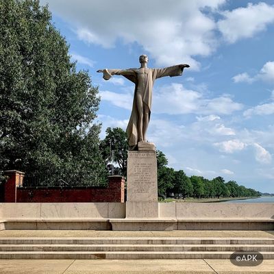 The Titanic Memorial statue in Washington, DC, depicting a robed figure with outstretched arms overlooking the waterfront, set against trees and an open sky.