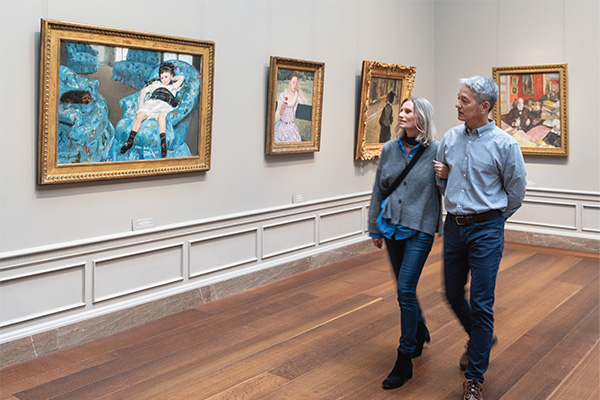 A couple walks arm in arm through a gallery at the National Gallery of Art, looking at framed paintings on a light-colored wall, including a large artwork featuring a reclining figure on a blue couch.