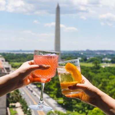 Two hands clinking cocktails at VUE rooftop bar with the Washington Monument and city views in the background.