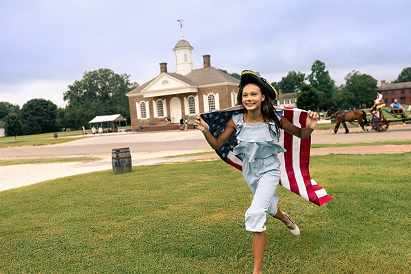 Child running on a grassy lawn at George Washington’s Mount Vernon, wearing a tricorn hat and holding an American flag, with the historic mansion in the background.
