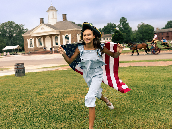 At a historic site in Colonial Williamsburg, a child wearing a colonial-era hat runs with an American flag draped across her shoulders. 