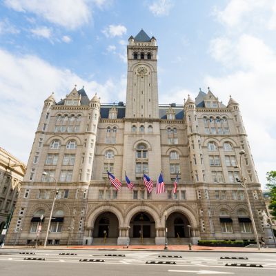Exterior view of the Waldorf Astoria Washington DC, featuring the historic Old Post Office building with its tall clock tower and American flags lining the entrance.
