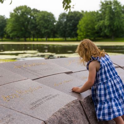 Child in a blue checkered dress exploring the engraved stone memorial at Signers Island, with a pond and trees in the background