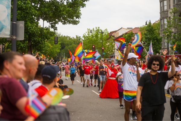 Crowds marching in a Pride parade carrying rainbow flags and wearing colorful outfits along a city street lined with spectators.
