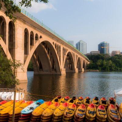 Colorful kayaks lined along the waterfront beneath Key Bridge, with the Potomac River and Rosslyn skyline in the background.