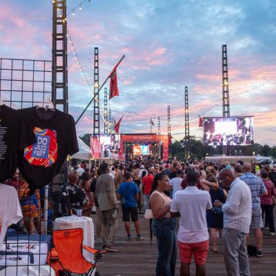 A large crowd gathers at an outdoor waterfront concert venue at sunset. A stage, video screens and festival merchandise are visible as attendees enjoy a live performance.