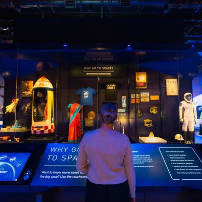 A visitor stands facing an interactive space exploration exhibit inside a dimly lit museum gallery. Displays include a model rocket, astronaut suit, artifacts and interpretive panels explaining the history and purpose of space travel.