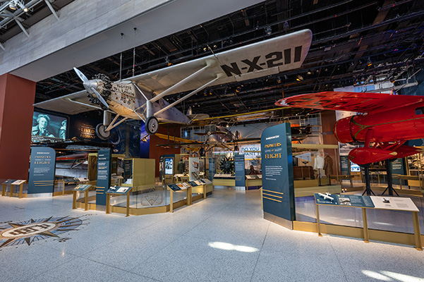 Historic museum gallery featuring a suspended vintage airplane above interactive exhibits on early aviation pioneers.