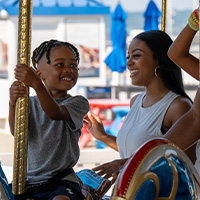 A young boy rides a carousel with his mother standing next to him.