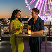 A man and a woman standing at a cocktail table with drinks at night. The Capital Wheel is brightly lit in the background.