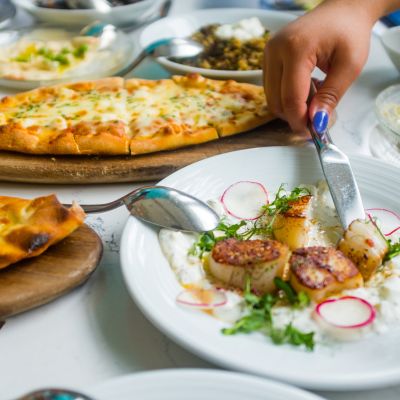 Person serving a plate of seared scallops beside flatbread and shared dishes on a restaurant table.