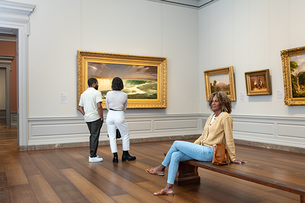 Visitors exploring a gallery at the National Gallery of Art, with one visitor seated on a bench while others admire framed landscape paintings on the walls.