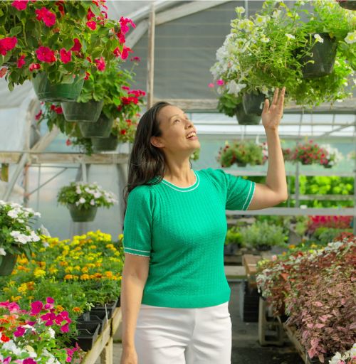 Smiling woman in a greenhouse reaching toward vibrant hanging flower baskets surrounded by blooming plants.