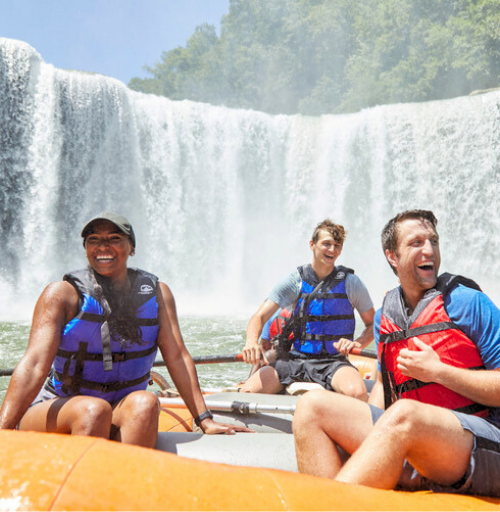 Three smiling adventurers sit in a bright yellow raft at the base of Cumberland Falls, with the powerful cascade rising behind them in the background.