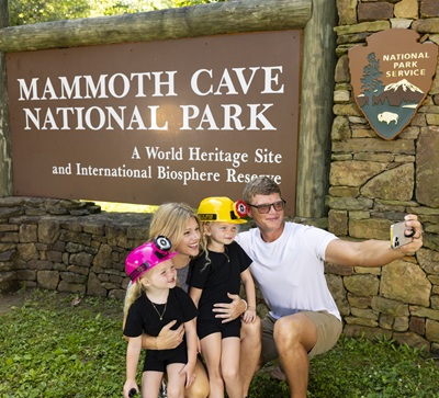A family poses in front of the Mammoth Cave National Park entrance sign while taking a selfie.