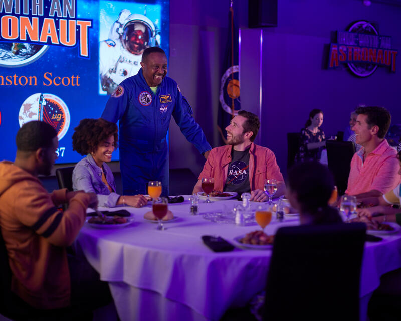 a man in a space suit standing next to a group of people sitting around a table
