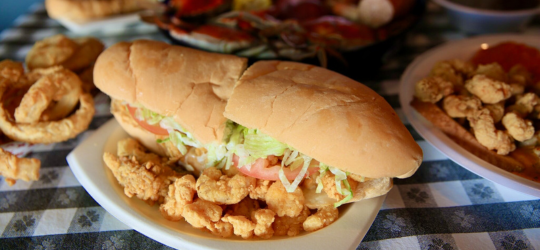 A close-up image of a fried shrimp po'boy, with various seafood items on the table behind it.