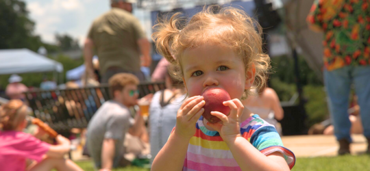 A little girl in a multi-colored dress takes a bite out of a peach at the Louisiana Peach Festival.