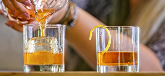 A closeup of a bartender pouring two sazerac cocktails.