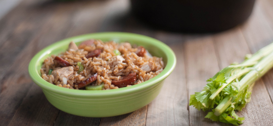 A bowl of chicken and sausage jambalaya rests on a table next to a stalk of celery.